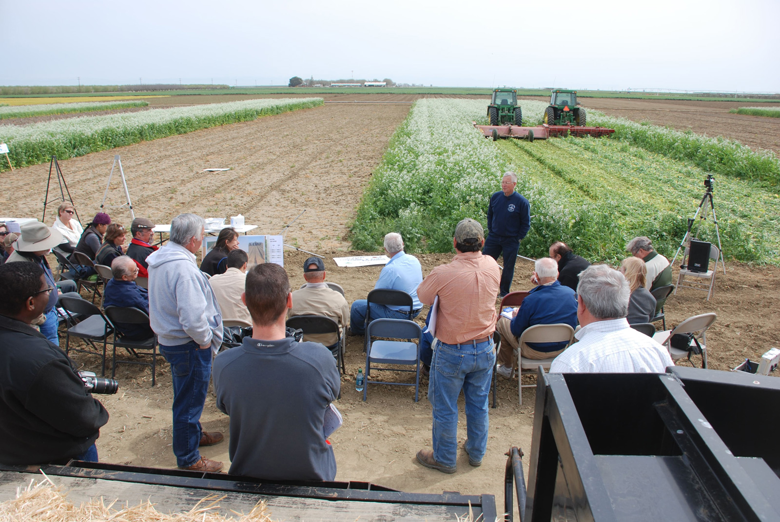 About 8 or 9 people standing outside by a field of green crops, listening to a man speaking.