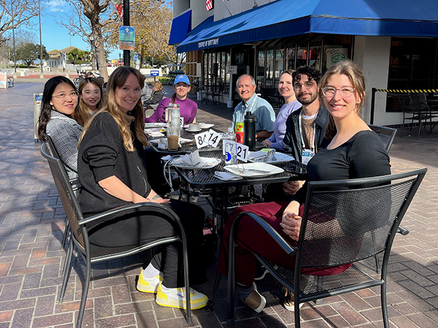 Seven young adults and an older man sitting at a table and smiling, outside on a sidewalk