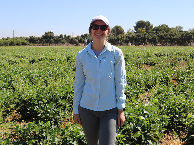 A young woman standing in a field of low-green, bushy plants, wearing casual clothes and a ballcap.
