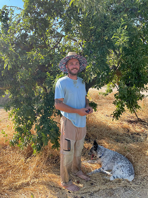 Young man wearing a hat, standing by a bushy tree. A spotted dog lays at his feet.