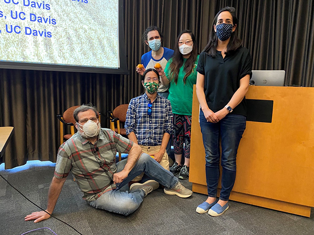 Several students standing near a podium in a curtained room. An older man lounges on the floor. They are making silly poses and wearing face masks.