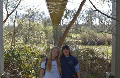 Two young women smiling under a bridge at a sunlit marsh with surrounding greenery. UC Davis Department of Plant Sciences