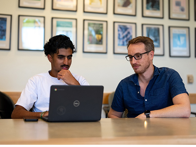 Two colleagues focused on a laptop at a table, framed photos on the wall behind