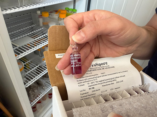 Hand holding small purple-capped  vial over open box in front of an open refrigerator in a scientific lab. UC Davis Department of Plant Sciences