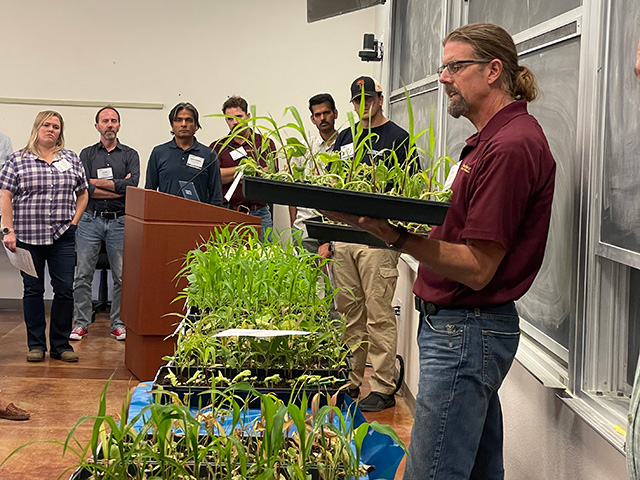 Man holding tray of green potted plants in classroom, people gathered along one side. UC Davis Department of Plant Sciences