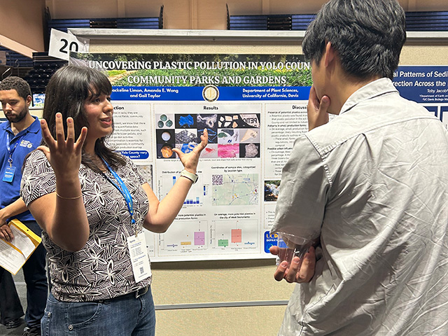 Young woman presenting in front of a conference poster, gesturing to a listener. UC Davis Department of Plant Sciences