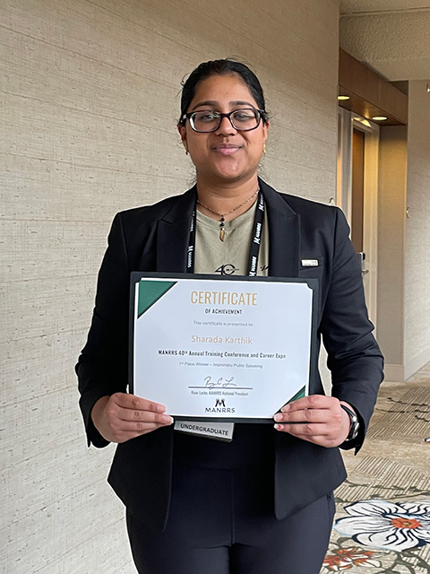 Young woman in dark blazer and glasses, smiles while holding a certificate in a hotel corridor. UC Davis Department of Plant Sciences