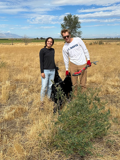 A young woman and young man in a wide-open rangeland, with a green busy plant in front of them. They are holding a large black plastic bag.