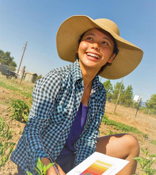 A smiling woman with a wide-brimmed hat sits in a field, holding a color palette. UC Davis Department of Plant Sciences