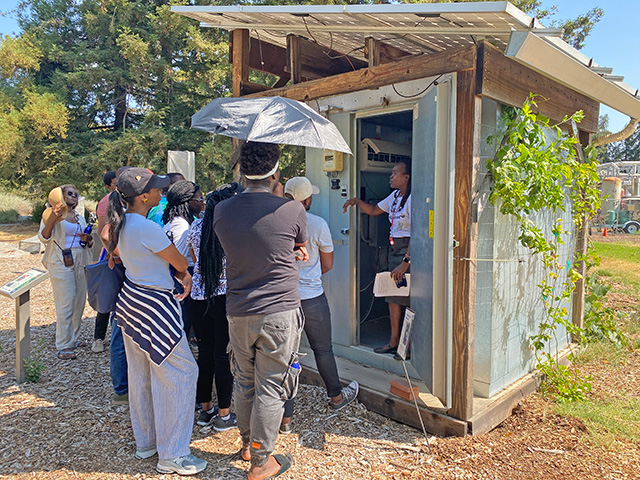 A small cinder-block structure. People stand around. A woman in the doorway speaks.