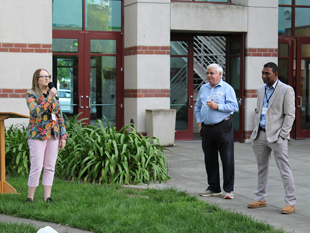 Three people standing outside a building. The woman is talking on a microphone