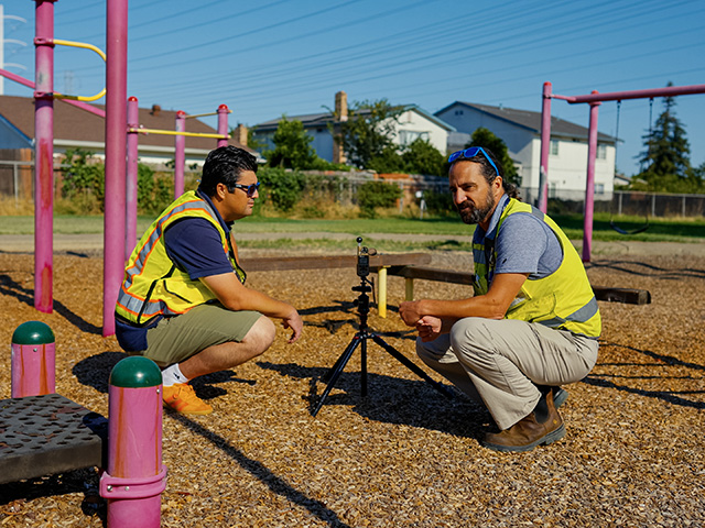 Two men wearing yellow safety vests squat in a school playground, talking. Blue sky overhead.