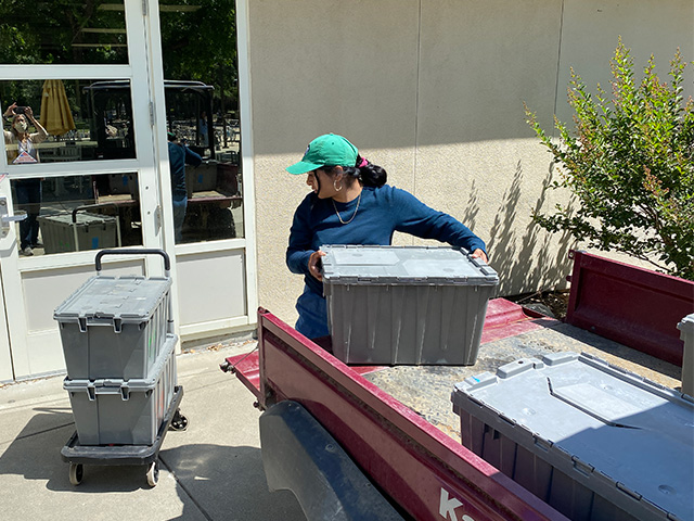 A woman outside a building lifts a large crate from a small truck onto the ground  near a door.