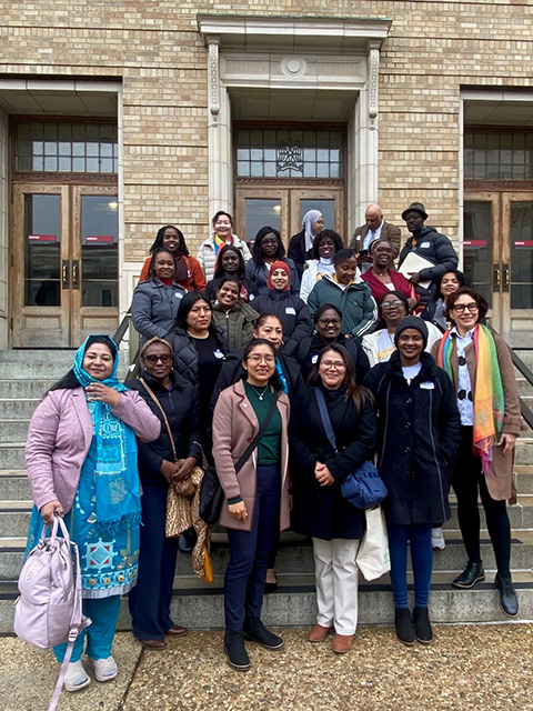 Group of about 15 women, many dressed in clothing from other countries, standing on the steps of an architecturally distinctive, pale-yellow-beige brick building.