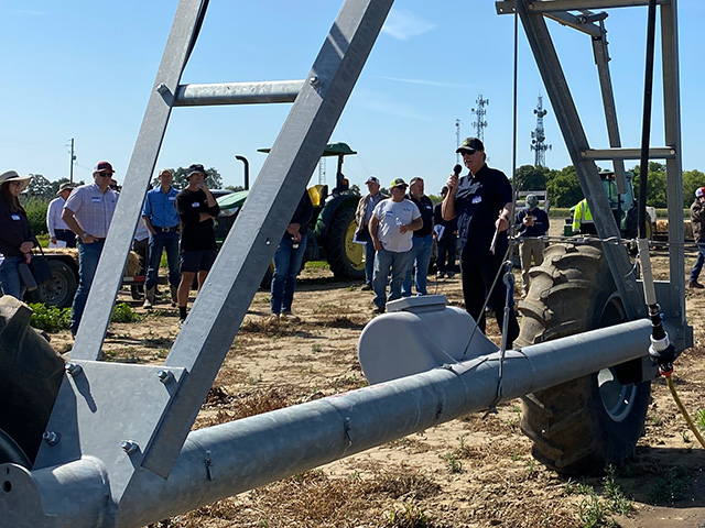 Close-up we see the geometric structure of a large metal apparatus. Inside the structure and farther beyond, a man speaks to a large group of people. They're out in a field of green plants.