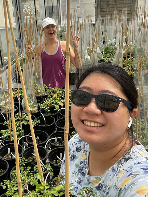 A woman close up in the foreground. Behind her are sticks poking up from trays of plants.