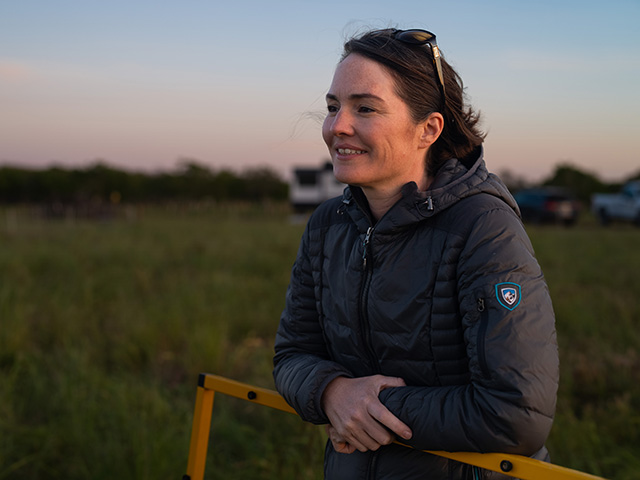 Woman standing outside in a field by a pasture fence, smiling. The sun is low and the light kind of orange.