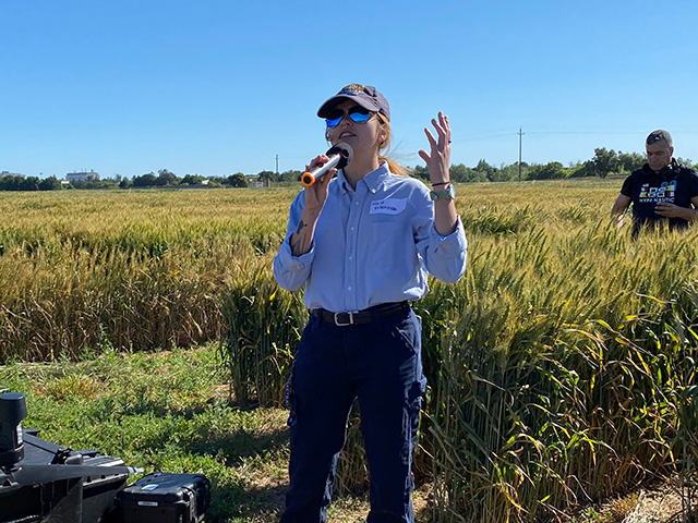 Women standing in front of tall, golden grain, holding a microphone. Blue sky above.