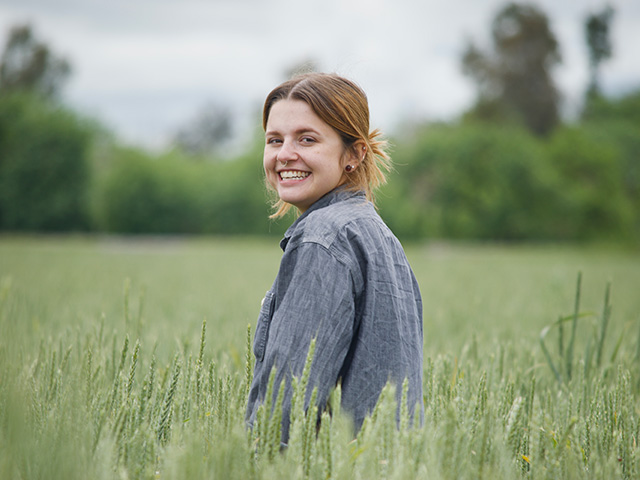 Smiling young woman looking back over her shoulder as she stands in a green field of grain under a cloudy sky. UC Davis Department of Plant Sciences