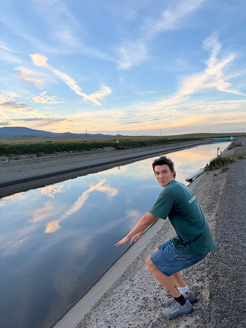 Young man crouching by canal at sunset, clouds reflecting in water