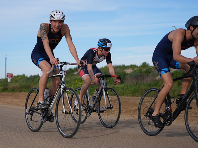 Three cyclists in helmets riding on a sunny rural road, focused expressions. The young man o the far right is grinning at the camera. UC Davis Department of Plant Sciencesat