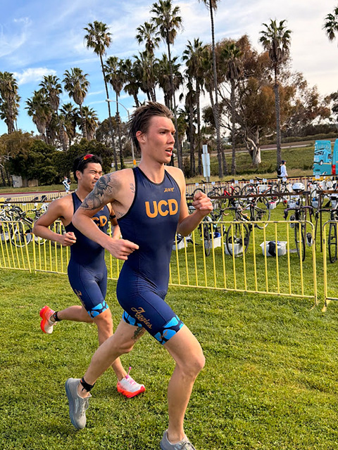 Two male triathletes running past racked bikes on grassy field, lead wearing UCD singlet.