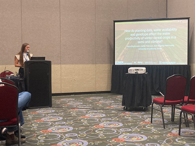 A young woman in a large room stands behind a podium, speaking, while a projection screen at a distance shows research findings