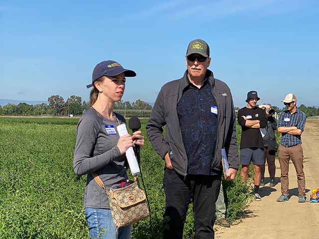 A woman and a man, standing in front of a field, with blue sky above.