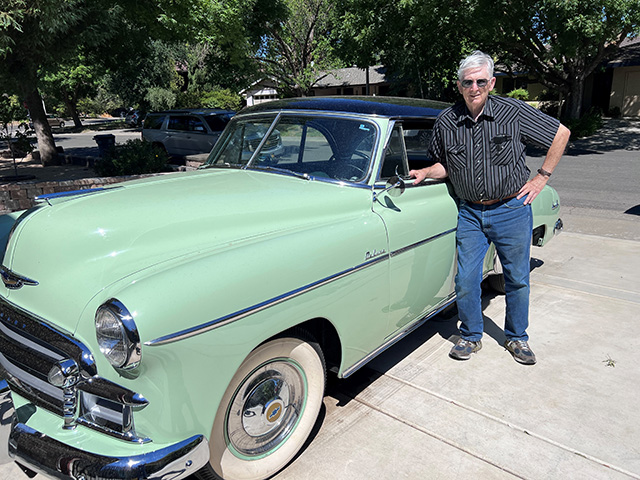 Older man standing next to mint-green vintage car on sunny suburban driveway
