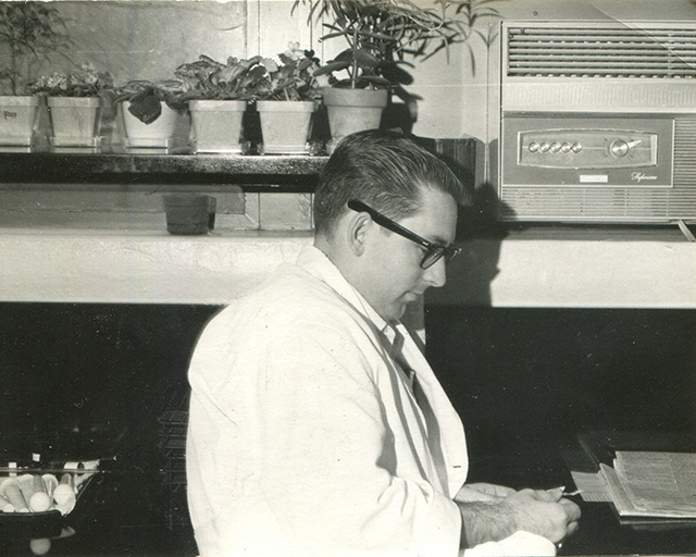 Black-and-white photo of a bespectacled man in a white coat writing at a desk with plants behind