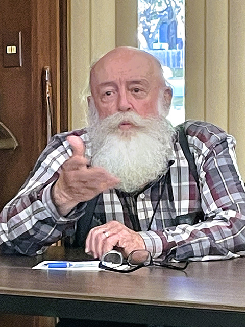A man with a long white beard and wearing a plaid shirt is seated at a table in front of a microphone