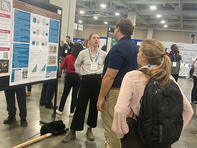 A young woman in a large room with lots of people. She stands by a research poster speaking, while a man and a woman look on.