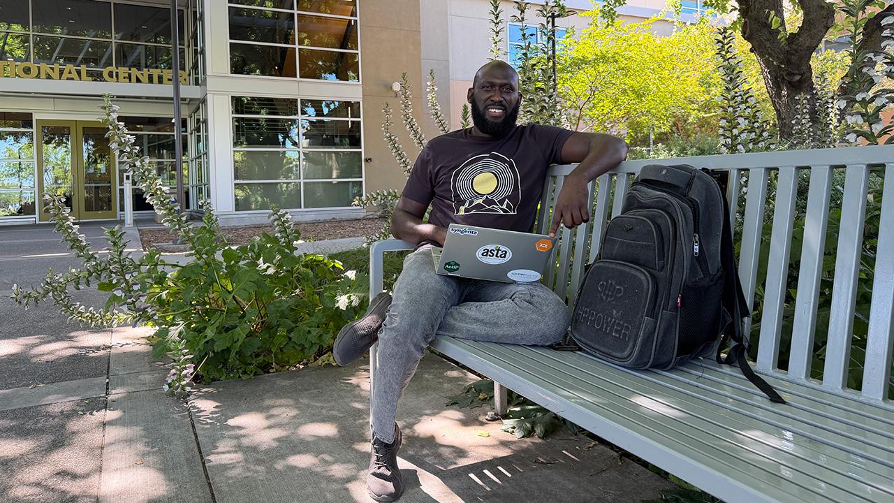 Man sitting on white bench in a garden, smiling with laptop on lap and backpack beside him.. UC Davis Department of Plant Sciences