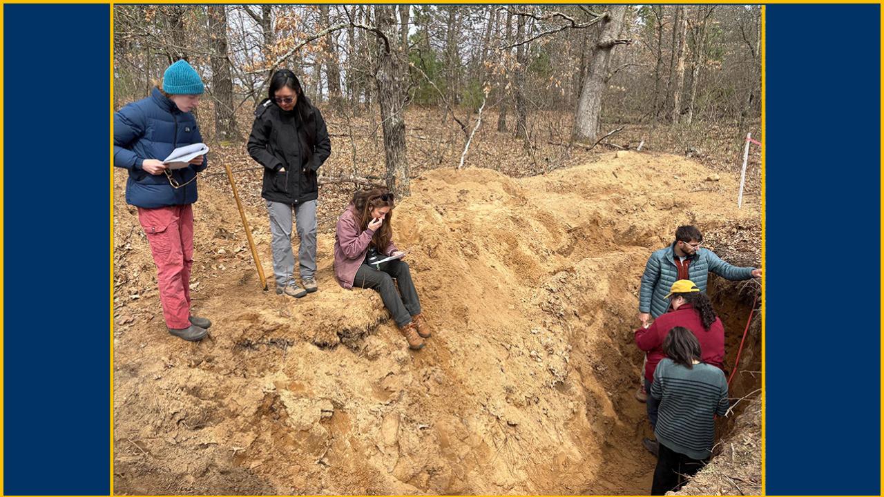 Three woman confer by the side of a pit dug by a forest. In the pit are three students.