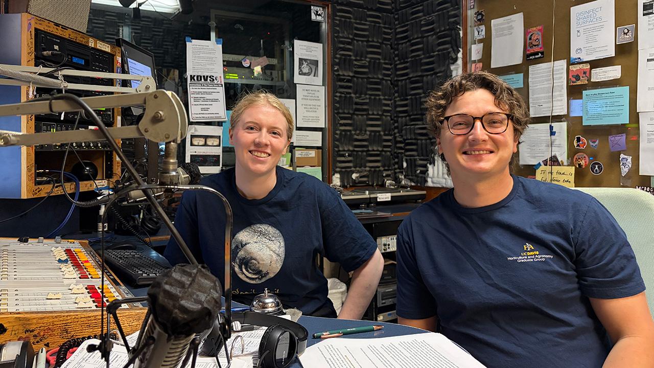 Two smiling people in radio studio with microphones and soundboard.