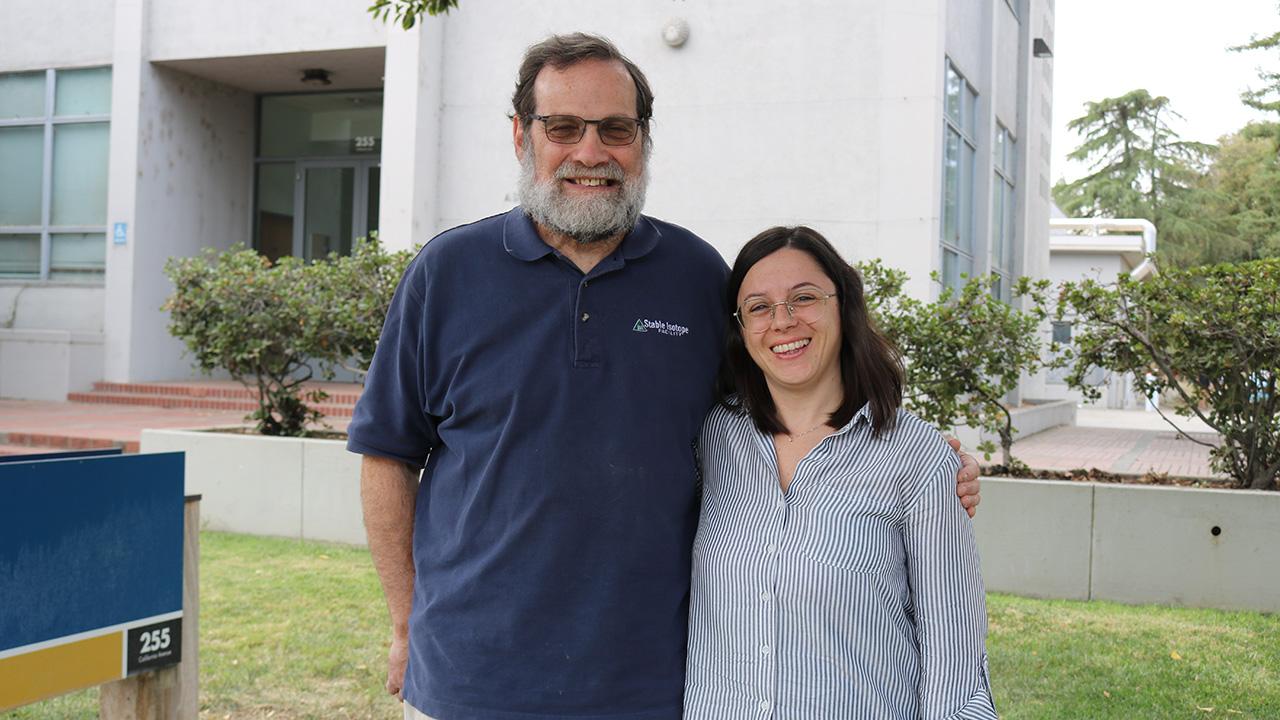 A man and a woman standing in front of a building. Green plants nearby