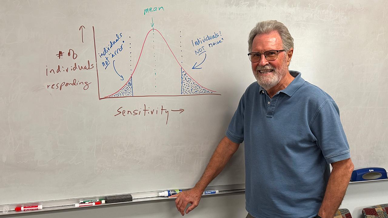 Smiling older man in blue polo standing by whiteboard with bell-curve graph. Notes on the board point arrows toward each end of the bell curve, saying "Individuals, NOT noise!" UC Davis Department of Plant Sciences