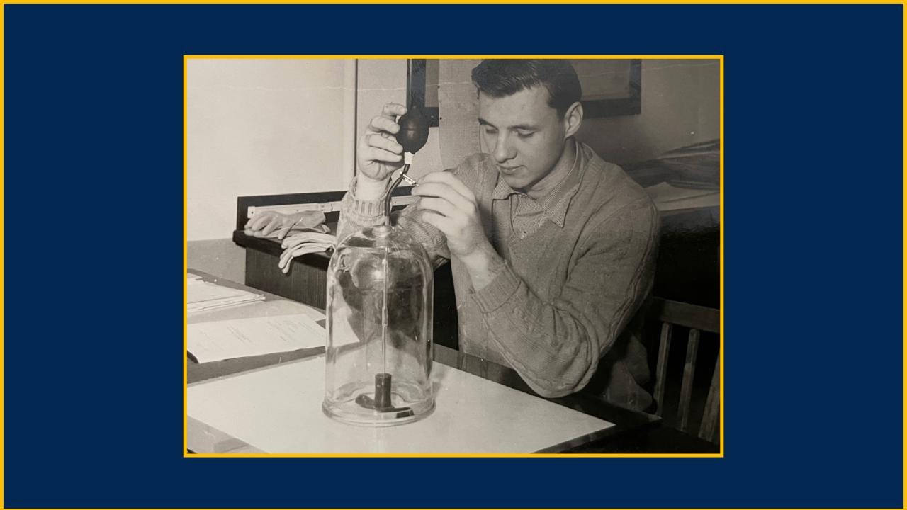 Black-and-white image of a young man in a lab setting with a glass beaker, doing something with it.