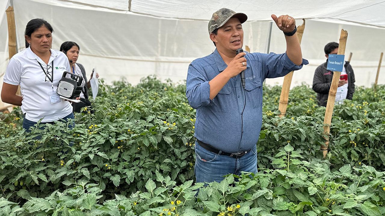 Man with a microphone speaking in a greenhouse-like structure, surrounded by green plants. A few people also visible in the background.