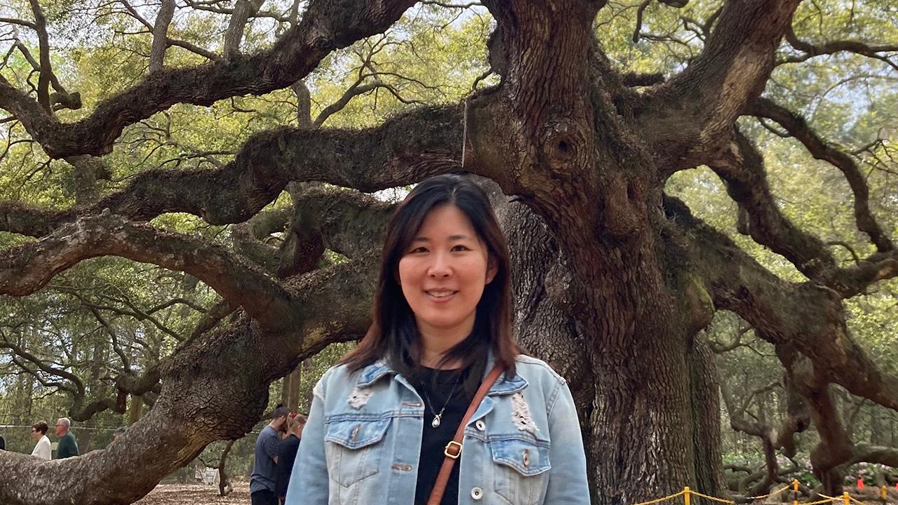 Smiling woman in denim jacket standing in front of a massive sprawling oak tree. UC Davis Department of Plant Sciences