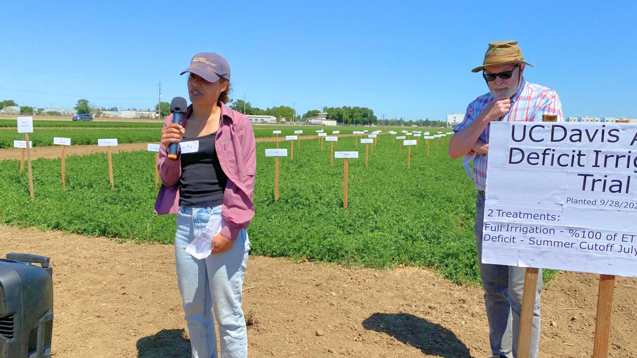 A young woman stands in front of a field, holding a microphone and speaking. 