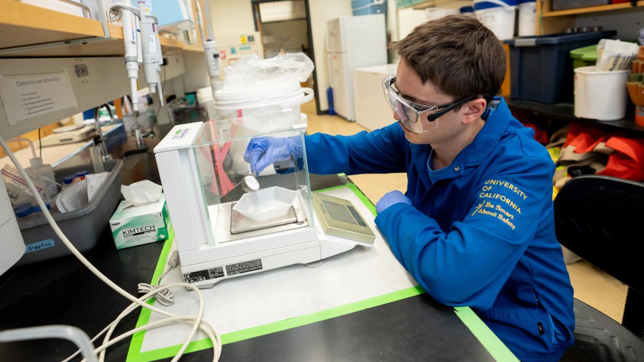 Young man in safety glasses using a lab balance at a chemistry bench. UC Davis Department of Plant Sciences