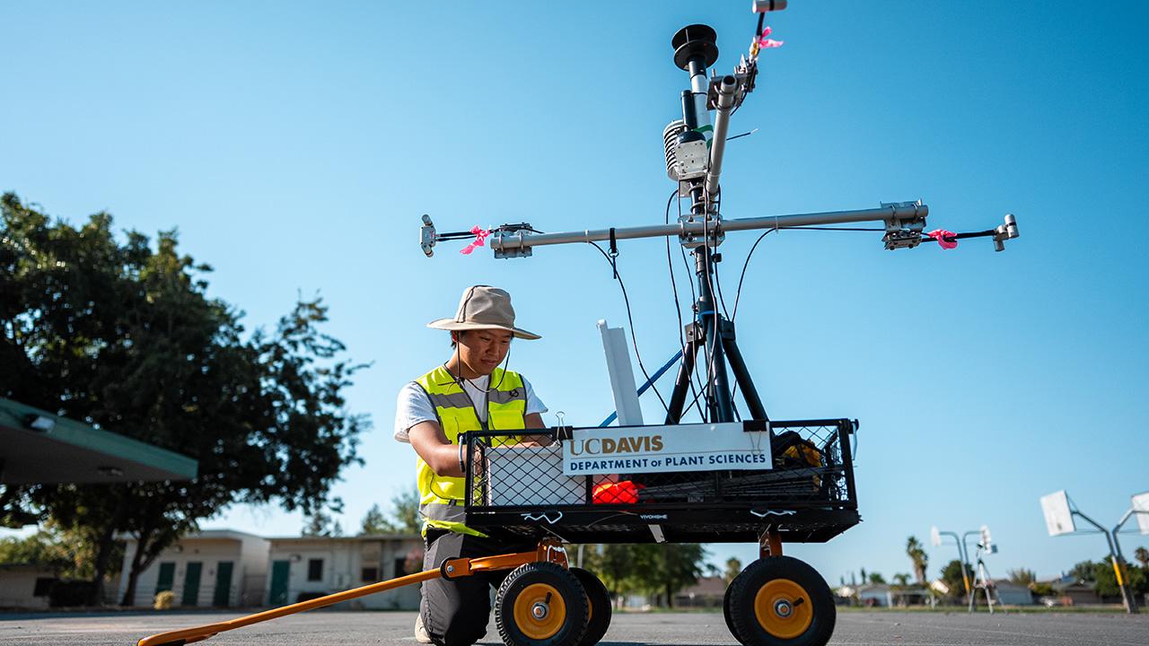 Young man wearing yellow safety vest and helmet kneels by a smallish wagon with a small tower on it. The tower has a cross-arm and more gear on it.
