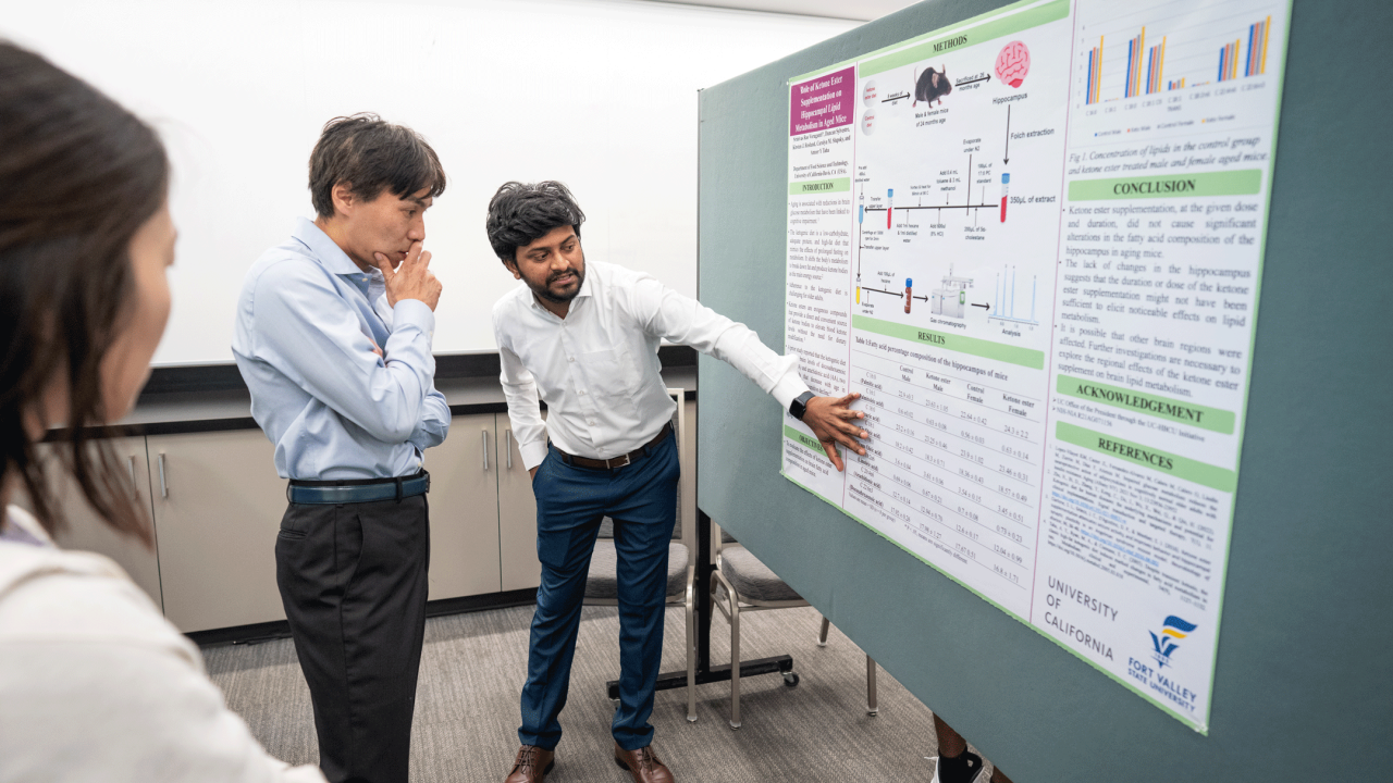 A younger man points to an academic poster, while an older man  looks on