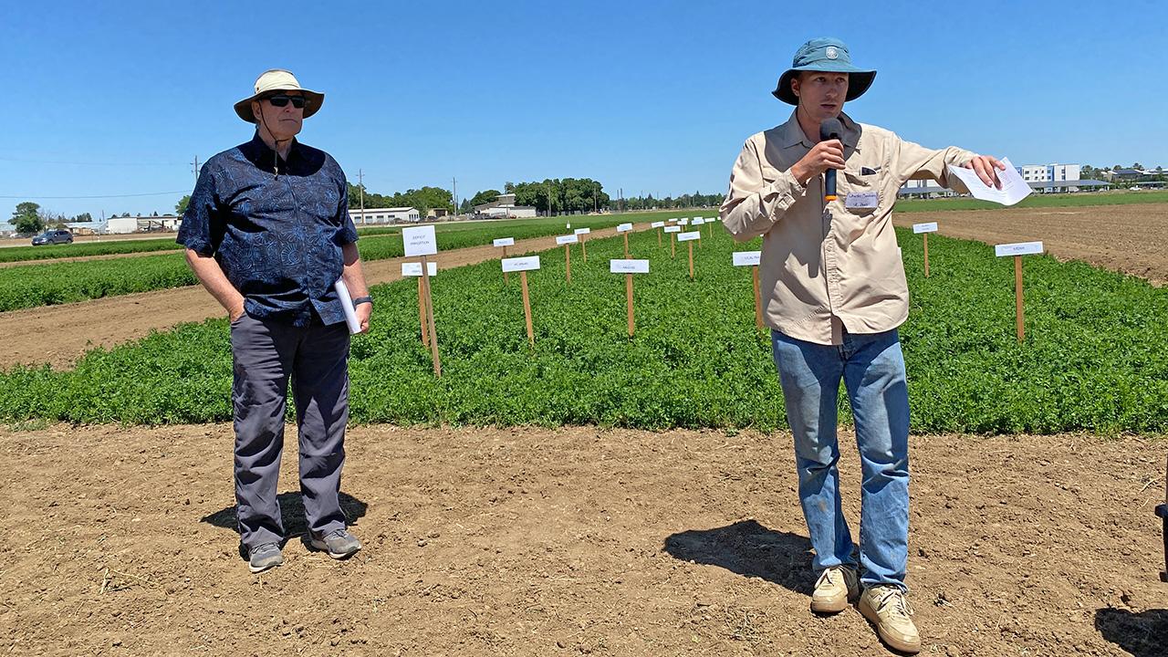An older man and a younger man in a field. The younger man has a microphone and is speaking and gesturing.