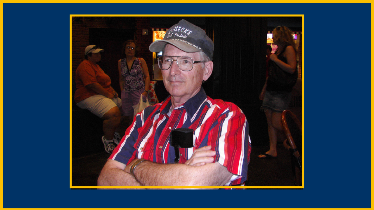 Older man in a ball cap and red-striped shirt, arms crossed, photographed at indoor social gathering