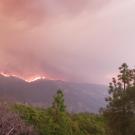 Orange-gray smoke billows above green trees, as flames rise from a mountain ridge.