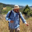 Man hiking through dry grass on sunny hillside, wearing hat, blue plaid shirt and backpack. Blue sky above and trees in background. UC Davis Department of Plant Sciences
