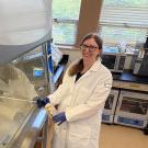 Woman in lab coat and gloves standing at biosafety cabinet with biohazard sticker, smiling. She holds a spray bottle in her hand. UC Davis Department of Plant Sciences