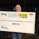 Woman smiling and holding oversized ceremonial check for €800 in dim indoor setting. UC Davis Department of Plant Sciences